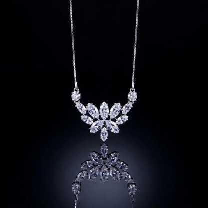 A detailed close-up product photograph of a delicate white-metal chain necklace hanging against a deep black, reflective background. The focus is on the central pendant, a sophisticated cluster of brilliant and marquise-cut shimmering stones carefully arranged into a symmetrical, multi-layered lotus or crown pattern. A partial, softened mirror reflection is visible directly beneath the pendant, enhancing its intricate sparkle. The chain is fine and unadorned.